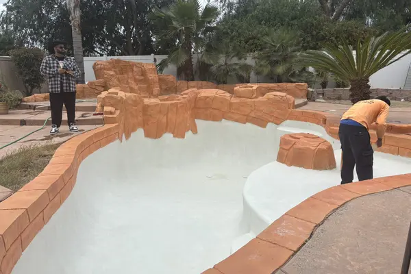 Vista a nivel del suelo de la piscina con enlucido blanco fresco, rocas esculpidas alrededor del borde y un trabajador terminando la superficie