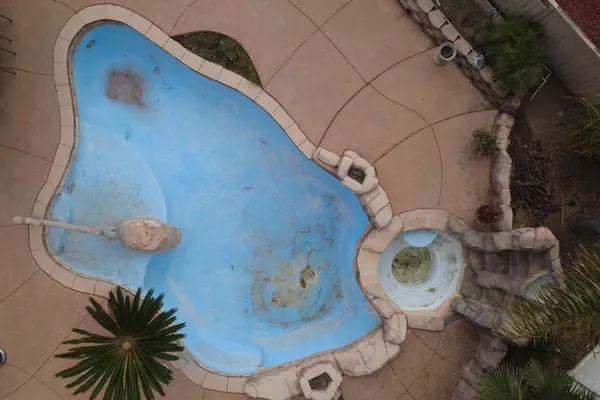 Vista aérea de una piscina de forma libre desgastada, con enlucido azul descolorido y manchas oscuras en todo el piso