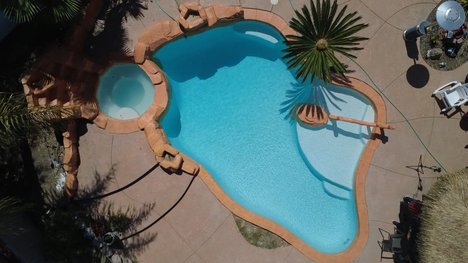 Vista aérea de una piscina de forma libre terminada, con cascada de roca y spa adjunto, llena de agua turquesa transparente